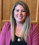 Ashley Conrad headshot in front of wood panel background wearing a rose colored blazer and black shirt.