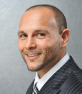 Headshot for John Vitale, wearing a grey pinstripe suit against a grey background