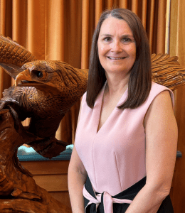 Melissa Gioglio stands next to a woodcarving of an eagle against a tan background. She is wearing a pink top.