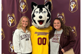 A Husky alum and admitted student stand on either side of mascot, Roongo in front of a maroon backdrop.