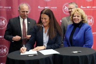 Four people standing behind a table. One person signing a paper. 