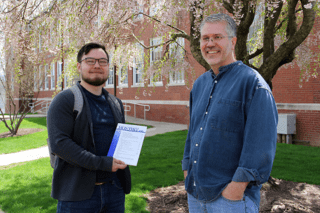 Two men standing next to each other outside. One of them is holding an award. 