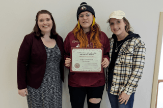 Three girls standing together. The middle one is holding an award. 