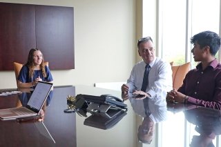 Three people sitting at a table with a phone and a laptop, talking. 