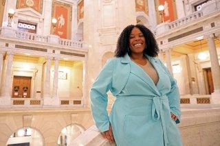 Brittany Stephenson, wearing a light blue coat over a tan shirt, standing in a large building with white pillars. 