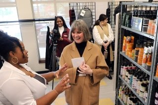 Two women talking to each other in front of a shelf. Someone in the background recording. 