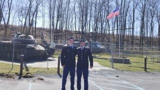 Two men standing in front of a fence and military equipment. 