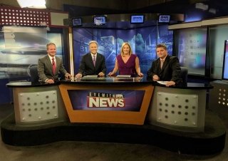 Four people sitting side by side at a news anchor desk. 