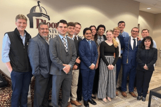 A group of 14 individuals dressed in business attire stand for a group photo in front of a beige colored wall with the Bloomsburg University Foundation logo mounted to the wall.