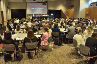 A wide angle photo of the Kehr Union ballroom filled with students at round tables, looking toward the front of the room where a projection on a screen displays the words "CATCH Conference"