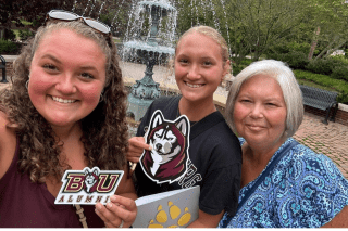 An alum and admitted student stand with their mom in front of the campus fountain