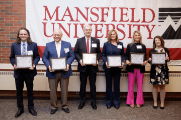 Six people standing side by side in front of a Mansfield University sign. They are each holding an award. 