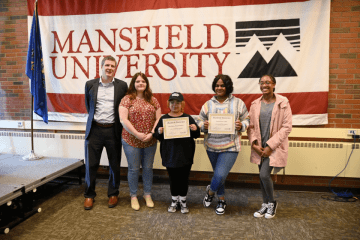 Five people standing side by side, in front of a Mansfield University sign. 