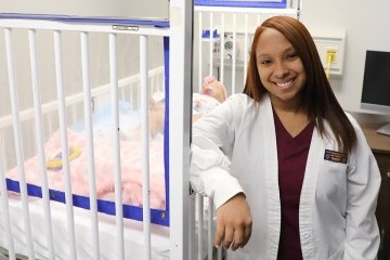 A nurse with a practice baby dummy in a crib behind her. 