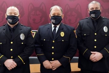 Three university police standing side by side with a red wall behind them. 