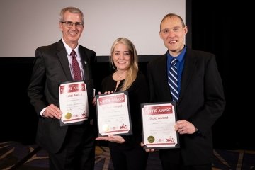 Three people standing side by side. They each have an award. 