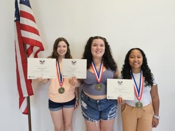 Three girls, with metals around their necks, standing next to an American flag, holding awards. 