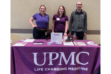 Three employees of UPMC stand behind a table draped by a purple tablecloth with white letters UPMC.