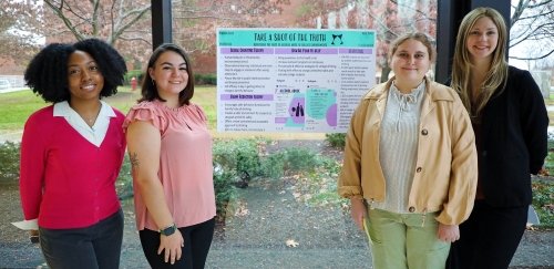 Students in front of health communications poster; 'Take a Shot of Truth' information relating to course studies