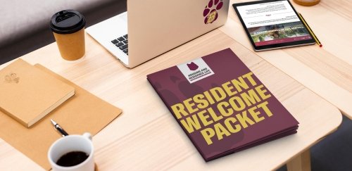 A laptop, iPad, coffee and Resident Welcome Packet folders on a desk.