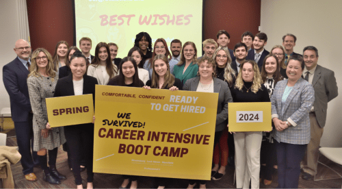 A group of students and staff members gather behind a yellow sign that says "We Survived CAREER INTENSIVE BOOT CAMP"