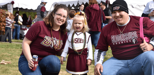 A Bloom alumni couple, wearing maroon BU shirts, crouch on either side of a little girl wearing a BU Huskies cheerleading outfit.