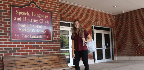 Audiology student standing outside Centennial Hall near the Speech, Language, and Hearing Clinic sign