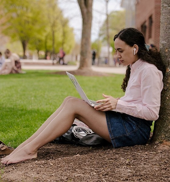 a student sitting under a tree in the quad working on her laptop