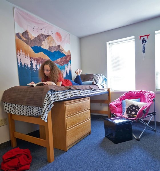 Female student laying in bed reading a book in a residence hall at CU-Mansfield