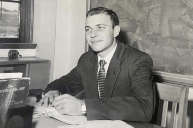 A black and white photo of a man, writing in a book, at a desk. 