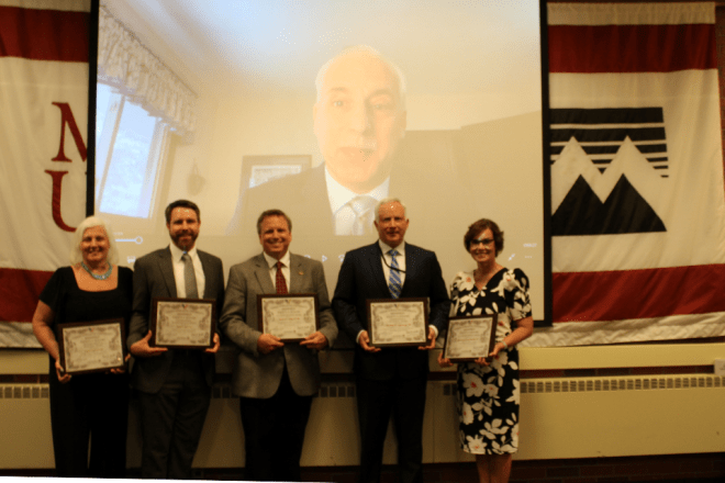 Five Mansfield alumni stand in front of a screen showing a sixth Mansfield alumnus. The five in-person alumni are each holding a plaque.