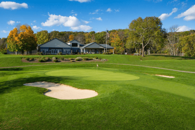 photo of a green and sand trap at Frosty Valley GC with the clubhouse and trees in the background against a blue sky.