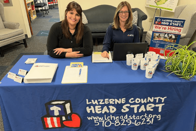 A photo of two women sitting at a table, wearing black henley collared shirts. The table has a blue tablecloth with the Luzerne County HEAD START logo and title.