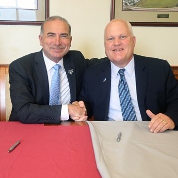 Two men sitting at a table. They are shaking hands. 