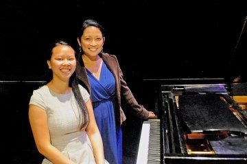 Two women standing with a piano. 