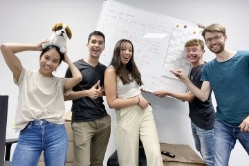 A group standing with a filled out whiteboard. One student with a stuffed dog on her head. 