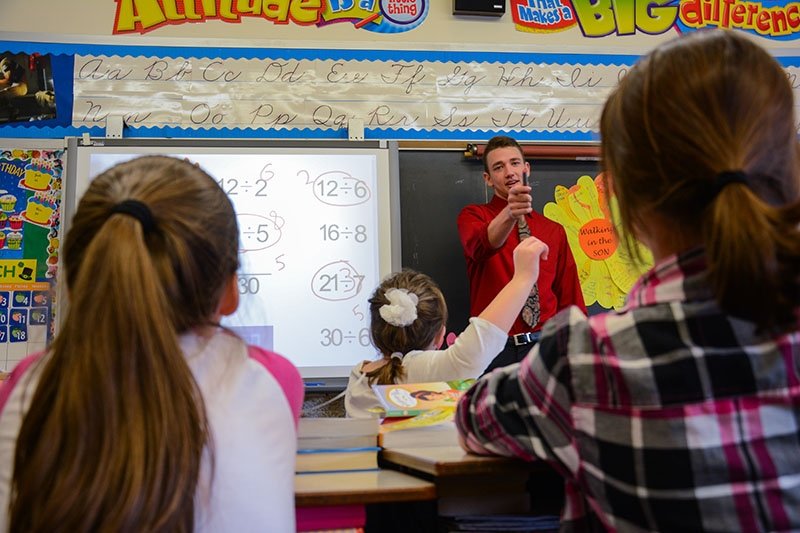 a teacher in front of a young class with a girl raising her hand to answer a math question