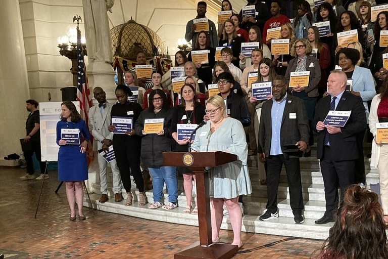 A group of people standing behind someone speaking at a podium. 