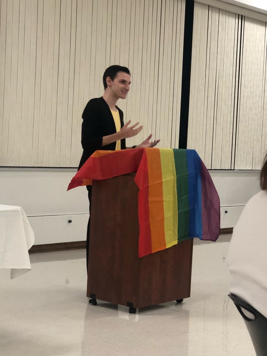 A person standing behind a podium with a rainbow flag laid across it. 